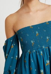 Teal off-the-shoulder-mekko, jossa on kukkakuviot, frilla ykkössivuilla ja valuvaa hihoja. Kangas on pehmeää ja rentoa istuvuutta.