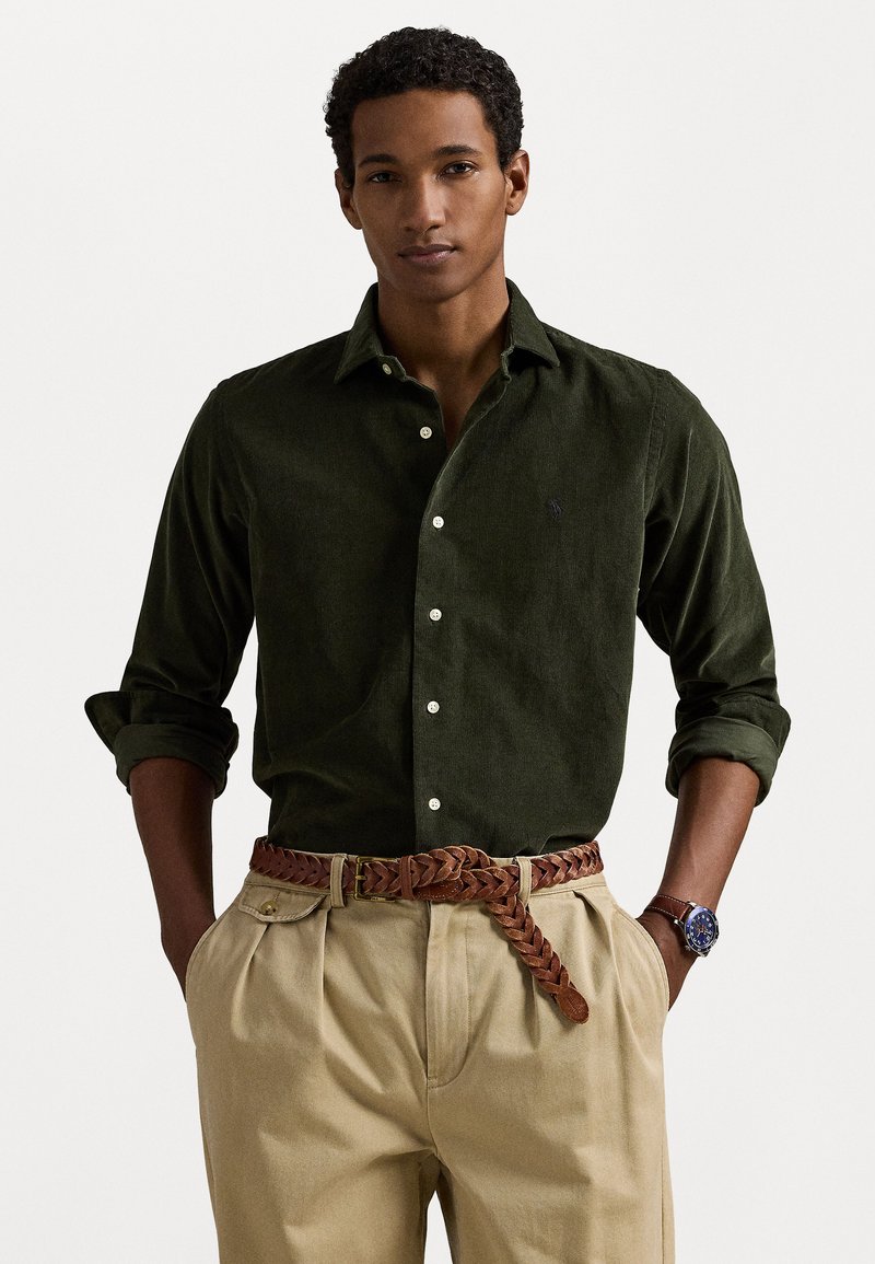 Chemise en velours côtelé vert foncé avec un devant à boutons et des manches retroussées, associée à un pantalon beige et une ceinture en cuir tressé marron.