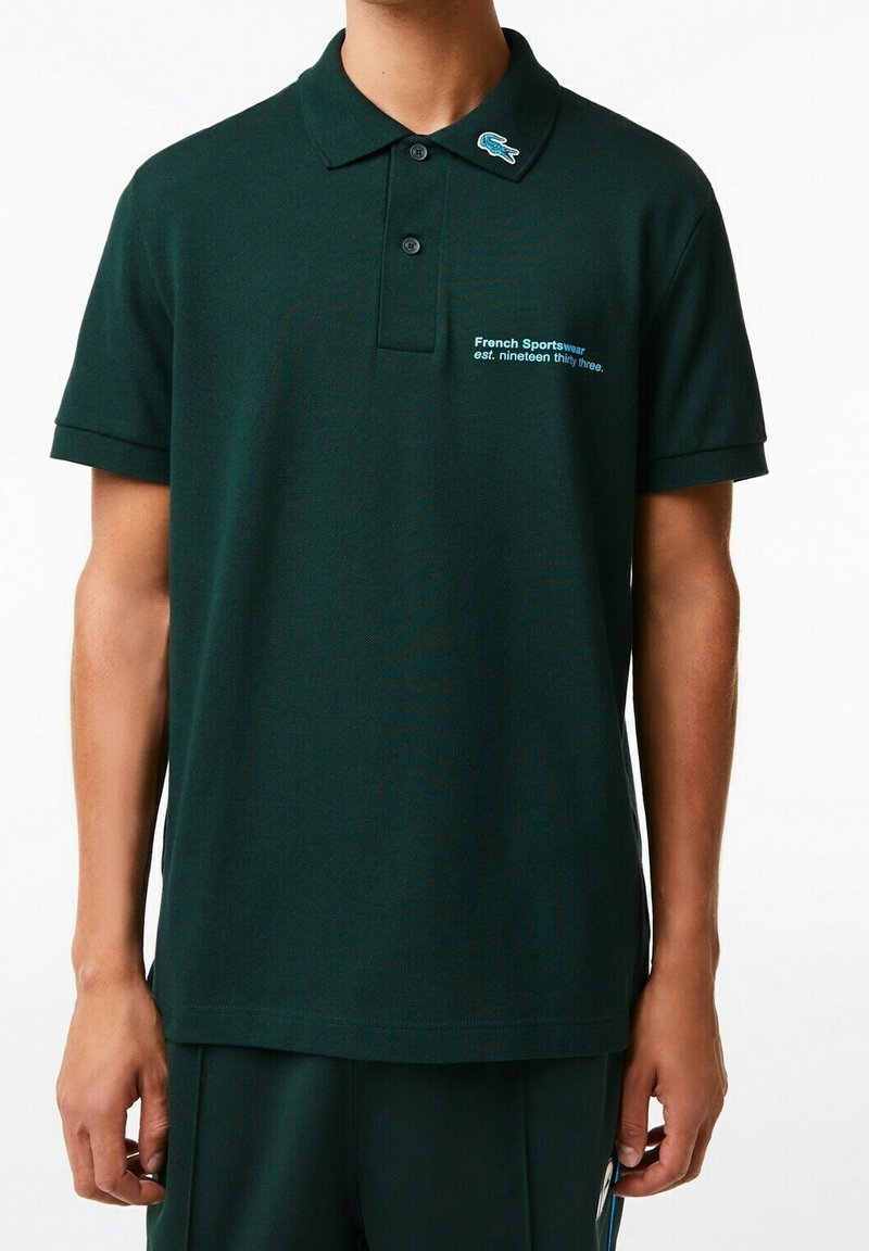 Polo-shirt vert foncé en tissu texturé, avec un plastron à boutons, des manches courtes et un logo brodé avec du texte blanc sur le devant.