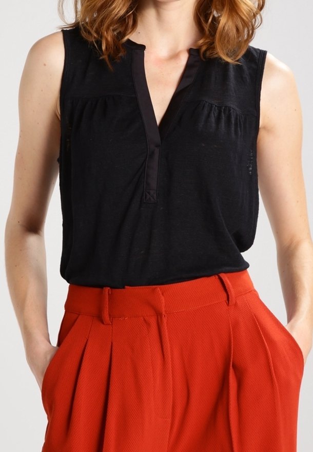 Femme portant un chemisier noir sans manches avec un col en V associé à un pantalon rouge texturé taille haute avec des plis et les mains dans les poches.
