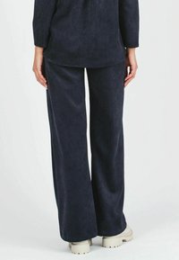 Pantaloni in corduroy blu navy con un design a gamba larga, caratterizzati da un tessuto testurizzato e una vestibilità rilassata, abbinati a calzature di colore chiaro.