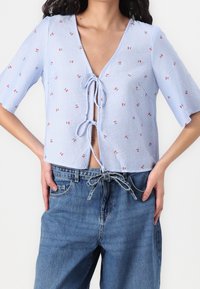 Blouse rayée bleue à manches courtes, avec motifs floraux et décolleté plongeant en V, nouée à l'avant avec un nœud ajustable. Portée avec un jean en denim.