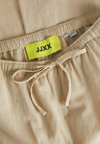 Pantaloni beige con vita elastica, lacci e un'etichetta gialla brillante con la scritta "JJXX." Il tessuto sembra morbido con una texture liscia.