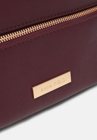 Bordowa torba z syntetycznego materiału z zamkiem błyskawicznym w złotym odcieniu oraz prostokątną złotą tabliczką z wytłoczonym nazwą marki. Teksturowana powierzchnia.