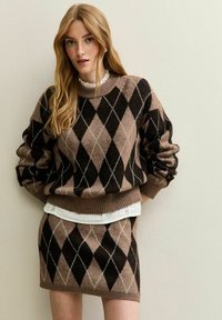 Femme aux longs cheveux blonds portant un pull en argyle marron et noir ainsi qu'une jupe assortie, debout devant un mur clair et uni.