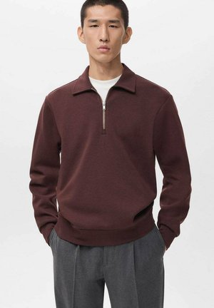 Felpa zip-up bordeaux con collo a camicia, caratterizzata da una cerniera argentata e polsini a costine, indossata sopra una T-shirt bianca e pantaloni grigi.
