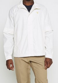 Mężczyzna w białej kurtce na zamek z złożonymi rękawami i beżowych spodniach, stojący na jednolitym jasnym tle.