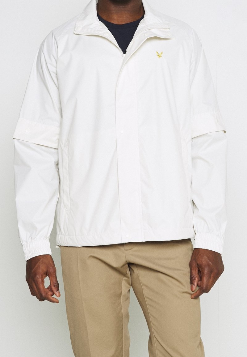 Mężczyzna w białej kurtce na zamek z złożonymi rękawami i beżowych spodniach, stojący na jednolitym jasnym tle.