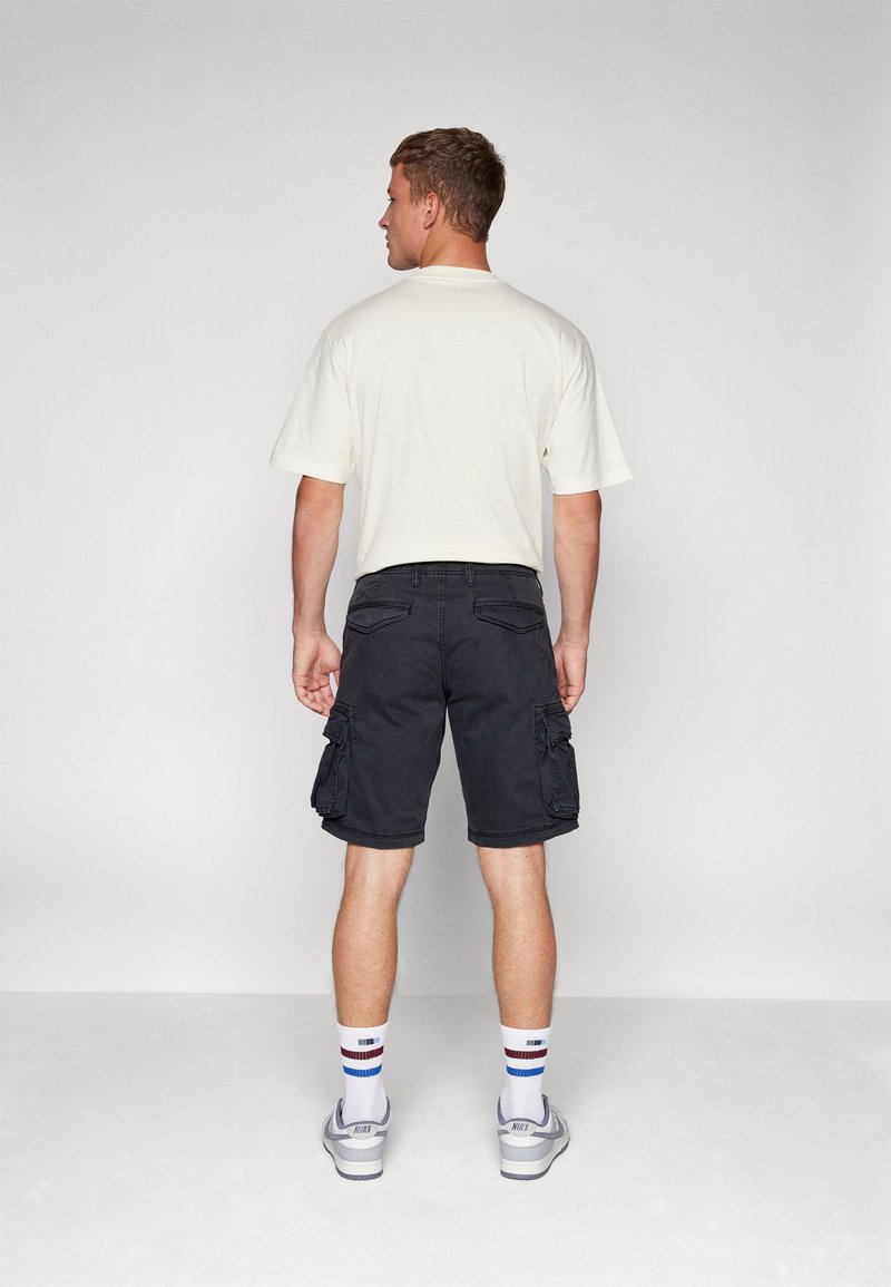 Pantalones cortos de carga negros con bolsillos laterales, combinados con una camiseta de color claro. Calcetines blancos con acentos azules y rojos, y zapatillas de deporte grises.