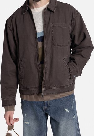 Mann trägt braune Jacke über gestreiftem Pullover und ausgewaschenen blauen Jeans, hält Sonnenbrille in der linken Hand vor weißem Hintergrund.