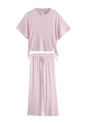 Fiatal nő hullámos barna hajjal, laza, világoslila rövid ujjú felsőt és hozzá illő húzózsinóros nadrágot visel, sima háttér előtt áll.