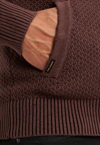 Brązowy sweter o teksturowanym wzorze, z prążkowanymi mankietami i dołem. Posiada boczną kieszeń z widoczną metką.