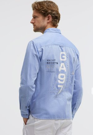 Man with short brown hair wearing a light blue long-sleeve shirt with yacht regatta text and sailboat graphic on the back.