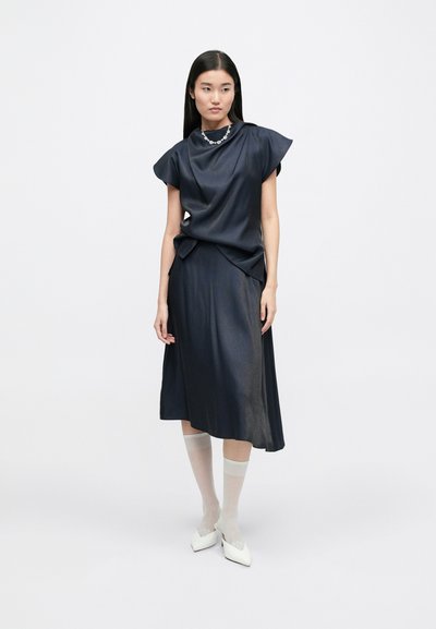 Navy satin dress with short, structured sleeves, draping detail at the waist, paired with sheer white stockings and pointed white shoes.