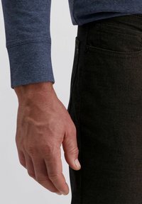 Mano maschile appoggiata accanto alla tasca dei jeans neri, indossando una camicia blu a manica lunga.