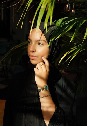 Hiyab negro y vestido con patrones intrincados, reloj de pulsera dorado y verde con correa texturizada, frente a un fondo de follaje verde.