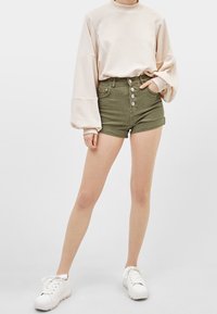 Ljusbeige långärmad topp med breda ärmar, kombinerad med olivgröna högmidjade shorts med fem knappar. Vita sneakers fulländar outfiten.