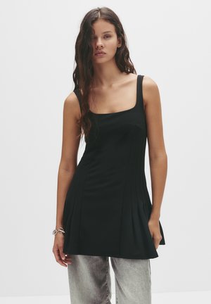 Tunica nera senza maniche con scollatura quadrata e design plissettato, realizzata in un tessuto morbido, abbinata a jeans grigi chiari.