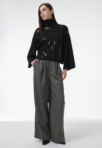 Schwarzes Oberteil aus strukturiertem Stoff mit goldenen Plattenakzenten, kombiniert mit grauen, weit geschnittenen Hosen im Hahnentrittmuster. Schwarze Absatzschuhe.