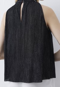 Černý top bez rukávů s vertikálními stříbrnými pruhy, vysoký límec s otvorem na knoflík na zádech, nošený osobou na bílém pozadí.