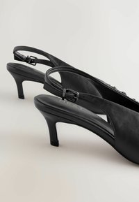 Schwarze Slingback-Absätze aus Leder mit schlankem Absatz und verstellbarem Schnallenriemen. Glatte Textur und minimalistisches Design. Auffälliges Logo auf der Innensohle.