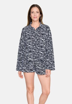 Femme debout portant un ensemble assorti de chemise à manches longues et short imprimés zèbre noir et blanc, sur un fond uni.