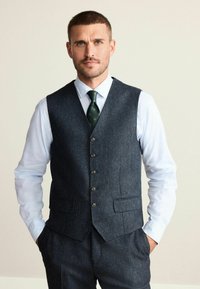 Mørkeblå tweedvest med fem knapper, to lommer, parret med en lyseblå skjorte og et grønt mønstret slips. Skræddersyet pasform.