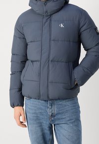 Muž oblečený v námořnicky modré péřovce s logy Calvin Klein na hrudi a rukávu, doplněné světlemodrými džíny.