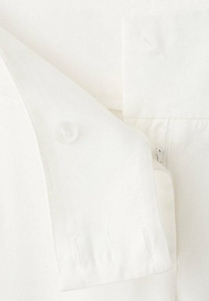 Gros plan des détails d’un vêtement blanc montrant un rabat boutonné et une fermeture éclair adjacente sur un tissu texturé.