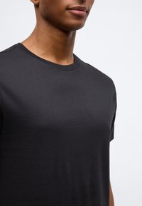 Prim-plan cu o persoană îmbrăcată cu un tricou simplu negru cu gât rotund, pe un fundal alb.