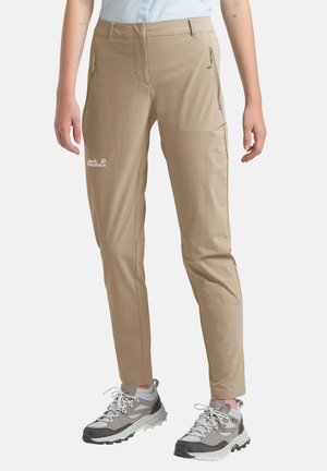 Beige Outdoor-Hose mit Reißverschlusstaschen, getragen mit grauen und weißen Wanderschuhen, Frontansicht von der Taille bis zu den Knöcheln.