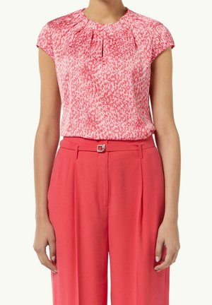Vrouw draagt een roze, met patroon, blouse met korte mouwen, ingestopt in een hooggetailleerde effen koraalkleurige broek met riem en zilveren gesp.