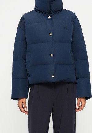 Marineblaue gesteppte Jacke mit Kapuze und goldenen Druckknöpfen, getragen zu schwarzen Hosen, zeigt den vorderen Oberkörper und die Hände.