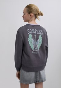 Gray sweatshirt featuring pastel-colored wings and a guitar graphic with "SCALPERS WORLD TOUR" text on the back. Cotton material, relaxed fit.