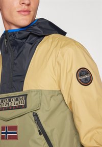 Farvet inddelt anorakjakke i beige, sort og olivengrøn. Har en lynlåslomme, logo-patch og justerbar hætte med blå kant.