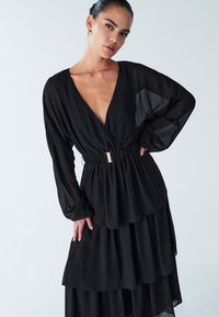 Frau mit zurückgekämmtem Haar, die ein mehrlagiges schwarzes Kleid mit tiefem V-Ausschnitt und tailliertem Gürtel trägt, posiert mit einer Hand in der Hüfte.