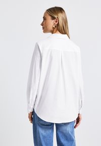 Chemise blanche à manches longues avec un ourlet arrondi et un pli au dos, associée à un jean en denim bleu clair. Le tissu semble lisse et léger.