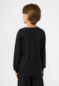 Enfant de dos portant un t-shirt noir à manches longues simple et un pantalon noir assorti, devant un fond clair et uni.