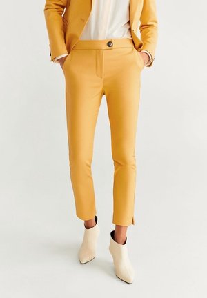 Vrouw met mosterdgele op maat gemaakte broek en bijpassend colbert, witte enkellaarzen, handen in zakken, tegen effen achtergrond.