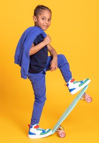 Dziecko w niebieskim stroju pozuje z deskorolką. Charakterystyczne cechy: kolorowe trampki za kostkę, teksturowana miętowa deskorolka, jednolite żółte tło.