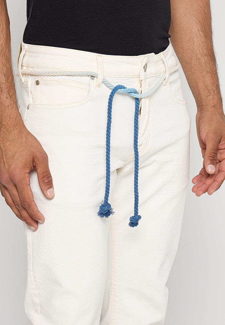 Pantalones blancos con un cinturón de cuerda azul, con textura trenzada y borlas, usados con una blusa negra; una mano descansa en la cadera.