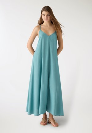 Giovane donna con lunghi capelli castani che indossa un abito lungo smanicato color turchese chiaro e sandali dorati, in piedi contro uno sfondo bianco semplice.