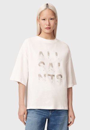 Jeune femme aux longs cheveux blonds portant un T-shirt blanc oversize avec le texte "ALL SAINTS" et un jean bleu, debout devant un fond uni.