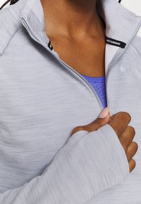 Valkoinen harmaa vetoketjullinen urheilujakku, joka on valmistettu sileästä kankaasta. Jakussa on korkea kaulus, sormet lämmittävät reiät ja hillitty tekstuuri. Musta logo näkyvissä.