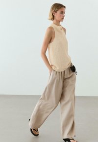 Frau, die geht, trägt ein ärmelloses beiges Strickoberteil, weite beige Hose, schwarze Sandalen und hält eine kleine schwarze Tasche an ihrer Seite.
