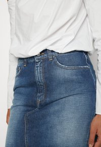 Femme portant une chemise blanche à manches longues rentrée dans une jupe en denim bleu délavé avec des poches avant et des coutures visibles.