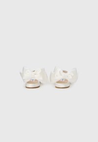 Λευκά σανδάλια σατέν με μεγάλα φιογκάκια στο πίσω μέρος. Τα λουριά είναι πλατιά με μπεζ σόλα και λεία υφή.