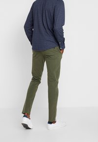 Oliivinvihreät, kapealahkeiset housut, joissa sileä rakenne, yhdistetty tummansiniseen nappipaitaan, jossa on pieni valkoinen kuvio, ja valkoisiin tennareihin.