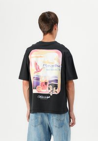 Zwarte katoenen T-shirt met een grote kleurrijke graphic van een vogel en een drankje op de achterkant, met de tekst "Paradiso Airlines" erboven.