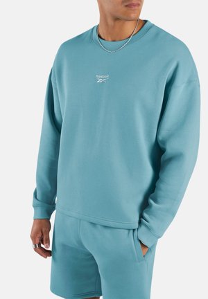 Mann trägt hellblauen Reebok-Sweatshirt und passende Shorts mit silberner Kettenhalskette und Ringen, steht vor einfachem Hintergrund.